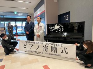 ピアノ贈呈式の横断幕とそれを持つ男女、ピアノの横に立つ男性２人