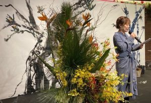 書道パフォーマンスステージ上、大きな生け花の横に司会の女性