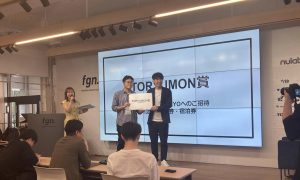 室内イベントの檀上に立つ二人の男性が賞状もちとなりに女性のMC