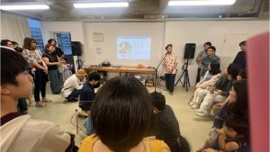 男性がパネルで事業説明。その周りに聴衆