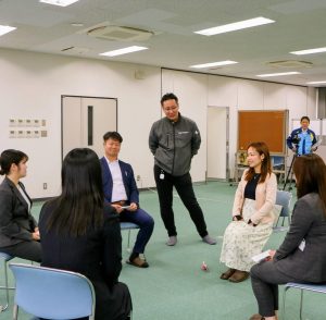丸く並べられた椅子に座り話をする女性４人男性１人、立って話す男性１人