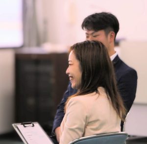 椅子に座って笑顔の女性、その奥に椅子に座る男性